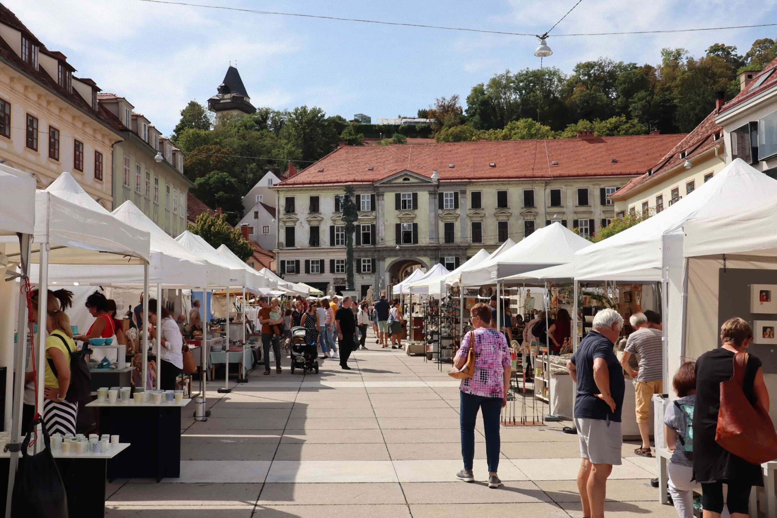 Besucher*innen zwischen den Marktständen mit Blick auf den Grazer Uhrturm.